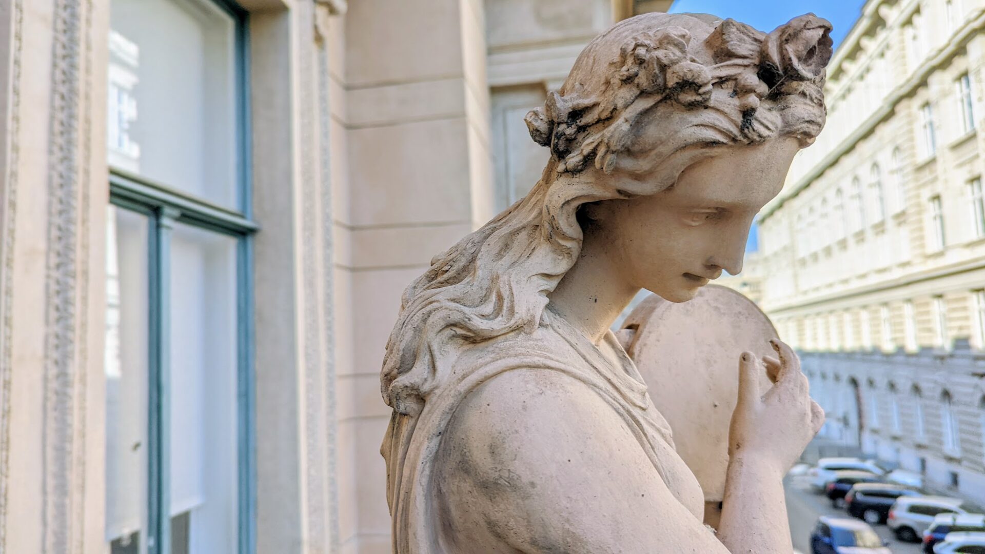 insolvence-Preston-vymahani-dluhopisu Marble statue of a woman with flowing hair in a city street, with buildings and parked cars in the background.