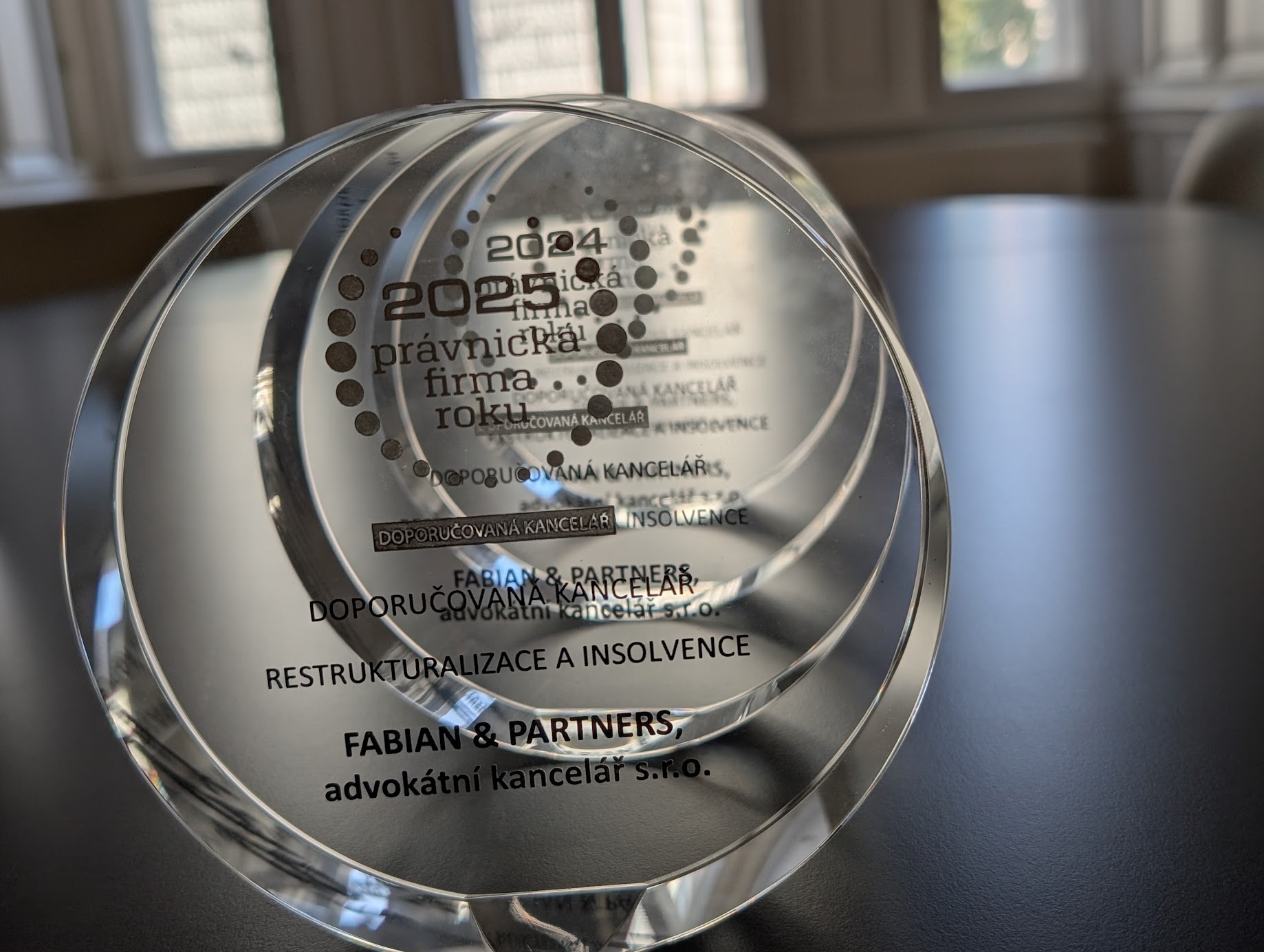 Insolvence Wood Seeds Moratorium Crystal circular award on a dark table, with Czech text and the name 'FABIAN & PARTNERS' visible, reflecting light through the glass.